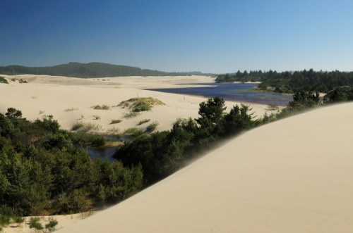 A serene landscape of sand dunes with green vegetation and a winding river under a clear blue sky.