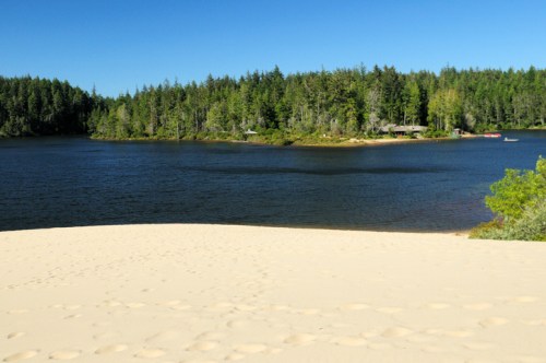 A sandy beach leads to a calm lake surrounded by lush green trees under a clear blue sky.