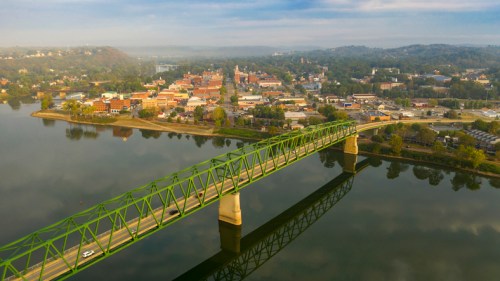Aerial view of a green bridge over a river, with a small town and hills in the background under a cloudy sky.
