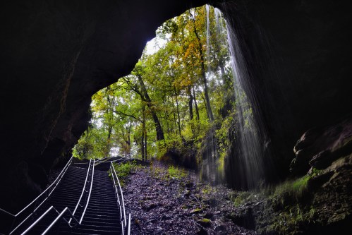 A staircase leads out of a cave, surrounded by lush greenery and a waterfall cascading down the rocks.