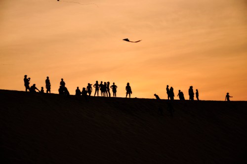 Silhouettes of people on a sand dune against a sunset, with a kite flying in the sky.