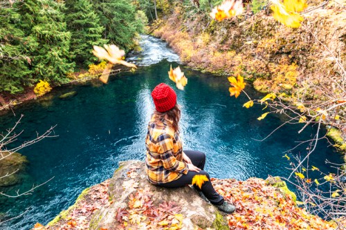 A person in a red hat sits on a rock by a river, surrounded by autumn leaves and lush greenery.