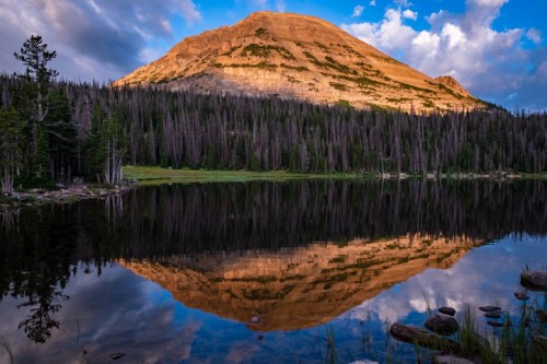 A mountain reflects in a calm lake, surrounded by trees and under a blue sky with scattered clouds.