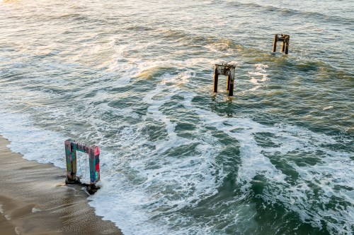 Three weathered pilings emerge from the ocean, partially submerged in waves, with colorful graffiti visible on their surfaces.