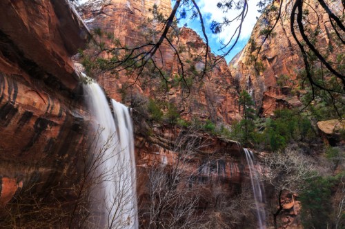 A scenic view of waterfalls cascading down red rock cliffs, surrounded by trees and a blue sky.