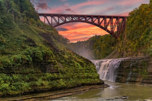A red bridge spans a lush green canyon with a waterfall, under a colorful sunset sky.
