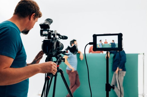 A filmmaker sets up a camera while a group of people poses in the background, with a monitor displaying the scene.