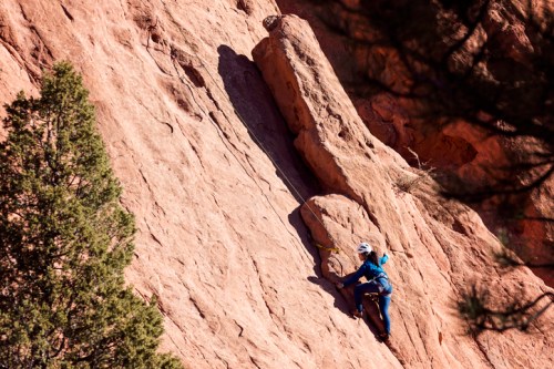 A climber ascends a rocky cliff, wearing a blue outfit and helmet, surrounded by trees and rugged terrain.