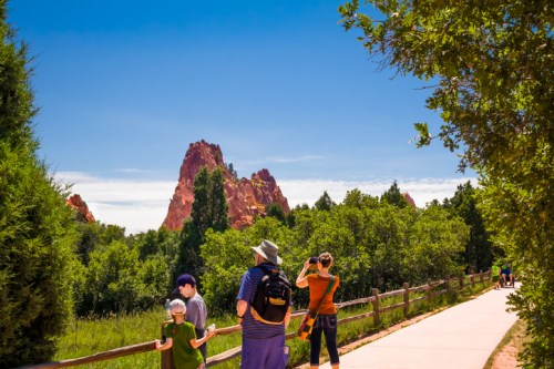 A family walks along a path with lush greenery, admiring a striking red rock formation under a clear blue sky.