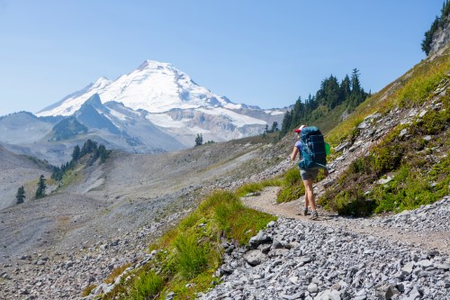 A hiker with a large backpack walks along a rocky trail, with a snow-capped mountain in the background under a clear blue sky.