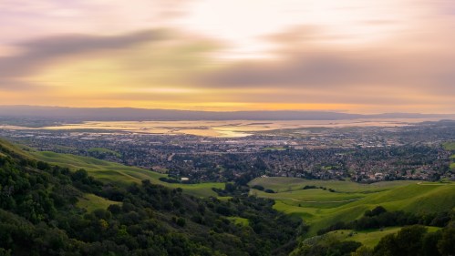 A panoramic view of rolling green hills and a shimmering bay at sunset, with soft clouds in the sky.