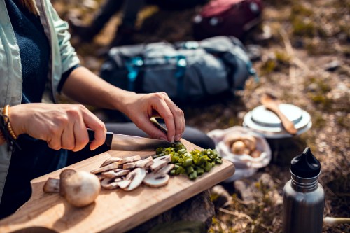 A person slices mushrooms and green onions on a wooden cutting board outdoors, with camping gear in the background.