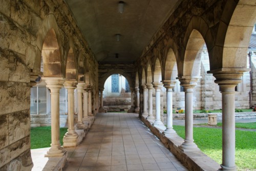 A stone corridor with arched columns, leading to a grassy area and a windowed wall in the background.