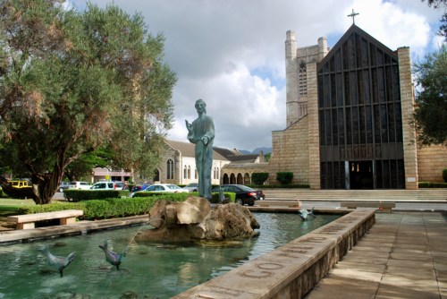 A statue of a figure holding a bird stands in a pond, with a modern church building in the background under cloudy skies.
