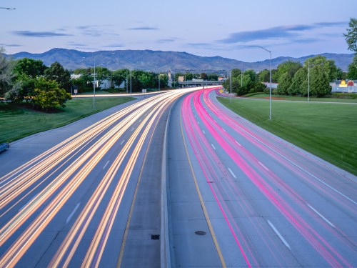 Light trails from vehicles on a highway, with mountains in the background and a clear blue sky at dusk.