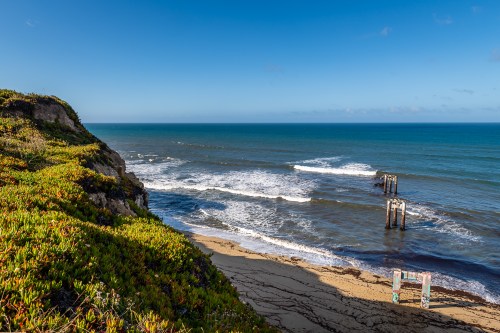 A scenic view of a beach with waves, cliffs, and remnants of a pier under a clear blue sky.