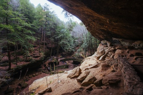 A rocky cave entrance overlooks a lush forested area with a small stream and a stone bridge in the distance.