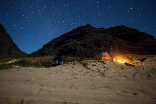A campsite on a sandy beach under a starry night sky, with a tent and a glowing campfire near mountains.