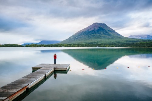 A person stands on a wooden dock by a calm lake, reflecting a mountain under a cloudy sky.