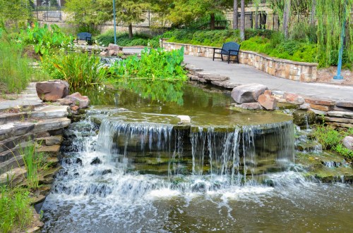 A serene park scene featuring a small waterfall flowing into a pond, surrounded by greenery and stone pathways.