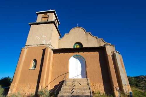 A weathered adobe church with a bell tower, featuring a white door and blue sky in the background.
