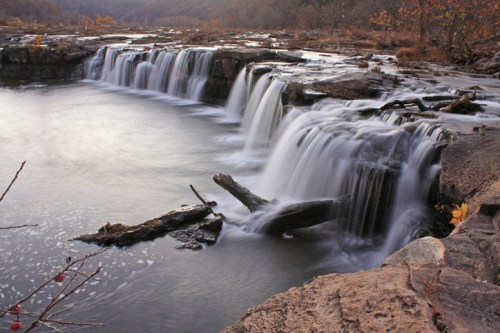 A serene waterfall cascades over rocks into a calm river, surrounded by autumn foliage and a tranquil landscape.