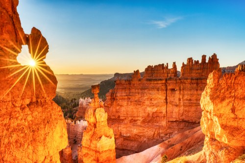 Sunrise over Bryce Canyon, highlighting unique rock formations and a starburst effect through a natural arch.