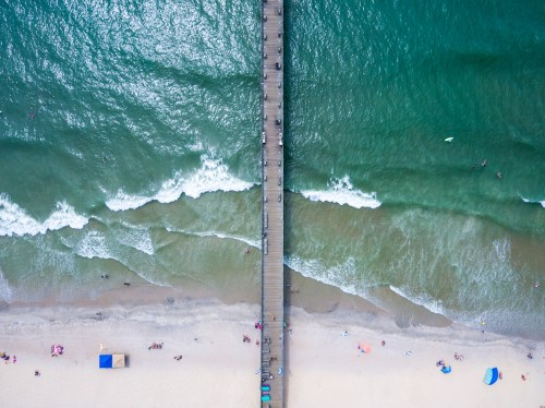 Aerial view of a beach with a wooden pier, waves, and people sunbathing on the sand.