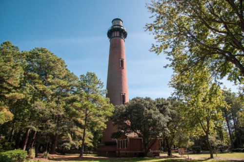 A tall brick lighthouse surrounded by trees under a clear blue sky.