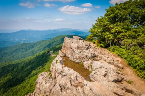 A rocky outcrop overlooks lush green mountains and a winding road under a blue sky with scattered clouds.