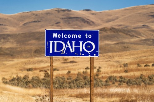 A blue welcome sign for Idaho stands in a dry, grassy landscape with rolling hills in the background.