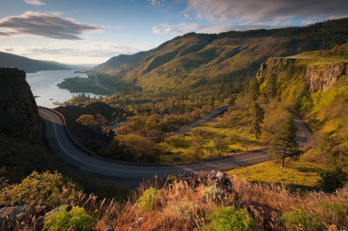 A winding road curves through lush green hills, overlooking a river and valley under a partly cloudy sky.