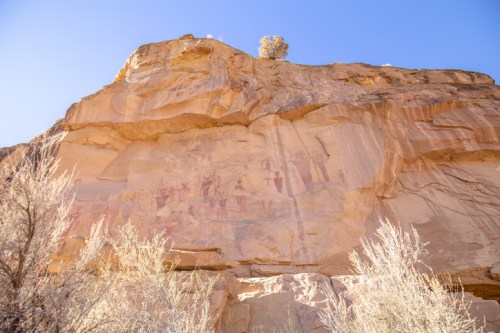 Ancient rock art on a cliffside, featuring faded figures and symbols against a clear blue sky.