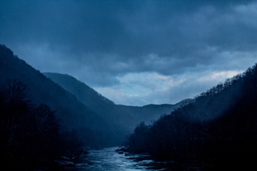 A dark, moody landscape featuring a river winding through misty mountains under a cloudy sky.
