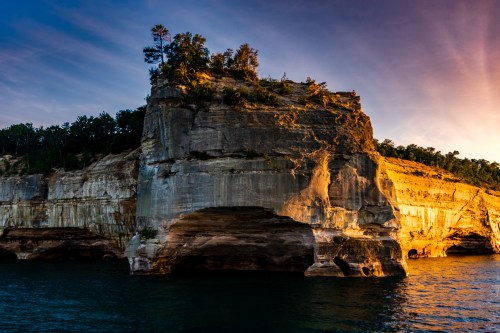 A rocky cliff with trees at the top, illuminated by sunset, rising above calm waters.