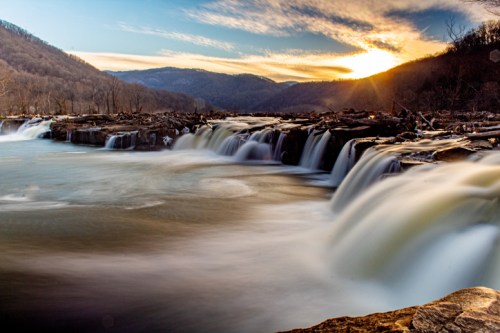A serene waterfall cascades over rocks at sunset, surrounded by mountains and a tranquil river.
