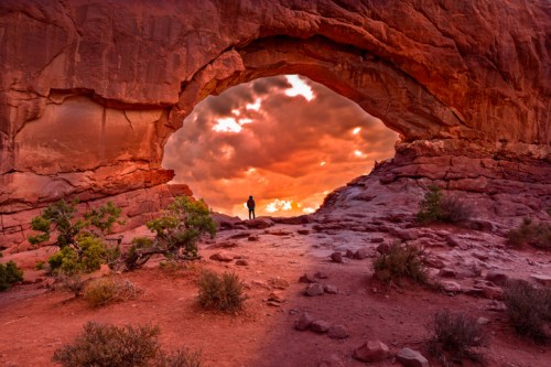 A person stands beneath a large rock arch, framed by vibrant sunset clouds and rugged red terrain.