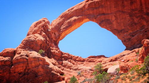 A natural rock arch stands against a clear blue sky, surrounded by red rock formations and sparse greenery.