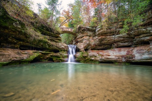 A serene waterfall cascades into a clear pool, surrounded by lush greenery and colorful autumn foliage.