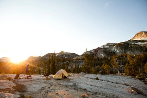 A person stands near a tent on rocky terrain, with mountains and a sunset in the background. Pine trees are scattered around.