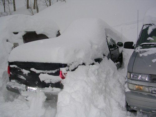 A car buried under heavy snow, with only the roof visible, surrounded by other snow-covered vehicles.