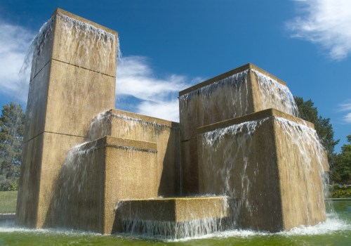 A modern stone fountain with cascading water, surrounded by greenery and a clear blue sky.