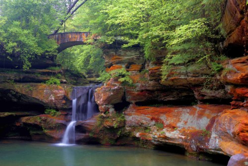 A serene waterfall cascades over colorful rock formations, with a stone bridge arching above in a lush green forest.