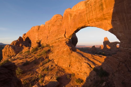 A natural rock arch stands against a sunset sky, surrounded by rugged terrain and desert vegetation.