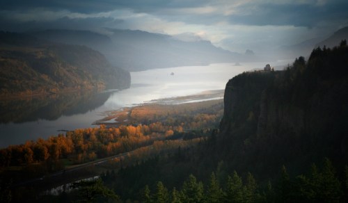 A misty landscape featuring a river winding through mountains, with autumn foliage and a distant cliffside structure.