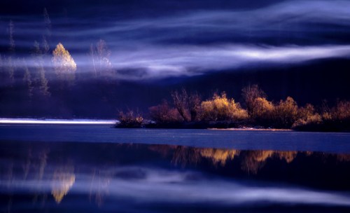 Misty lake scene with soft fog, reflecting trees and colorful foliage on the water's surface.