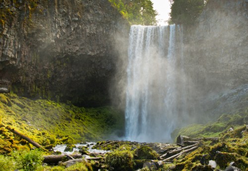 A majestic waterfall cascades down rocky cliffs, surrounded by lush greenery and mist rising from the water.
