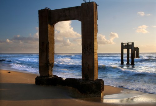 Weathered concrete pillars stand on a sandy beach, with waves gently lapping at their bases under a cloudy sky.