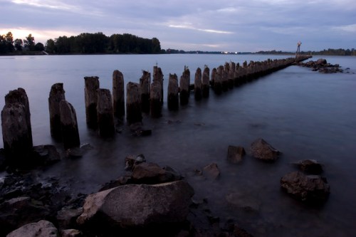 A calm river scene at dusk, featuring a weathered wooden pier extending into the water, surrounded by rocks and trees.