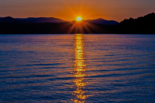A vibrant sunset over a calm lake, with golden rays reflecting on the water and mountains in the background.